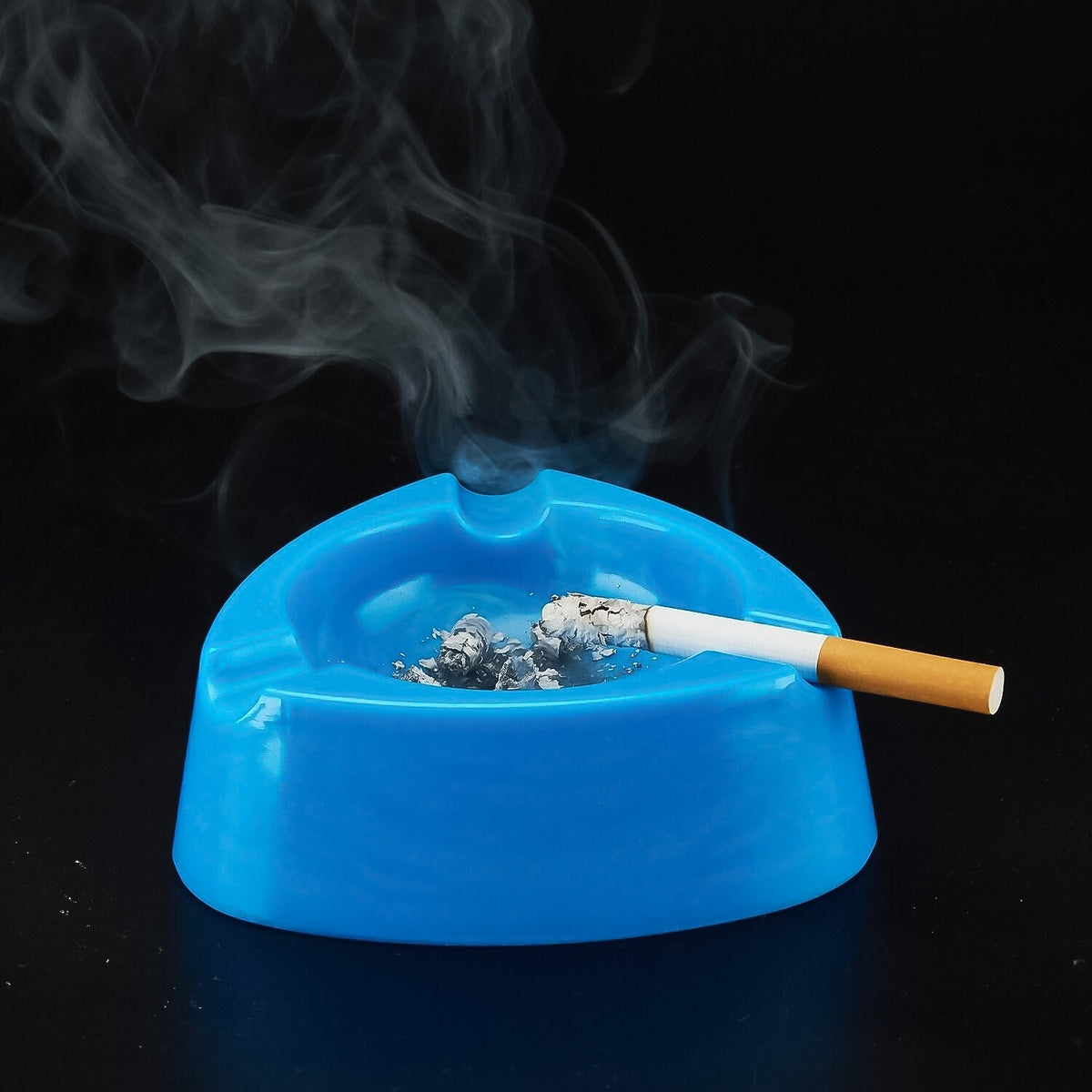 A lit cigarette rests in a blue plastic ashtray with ashes, emitting smoke against a dark background.