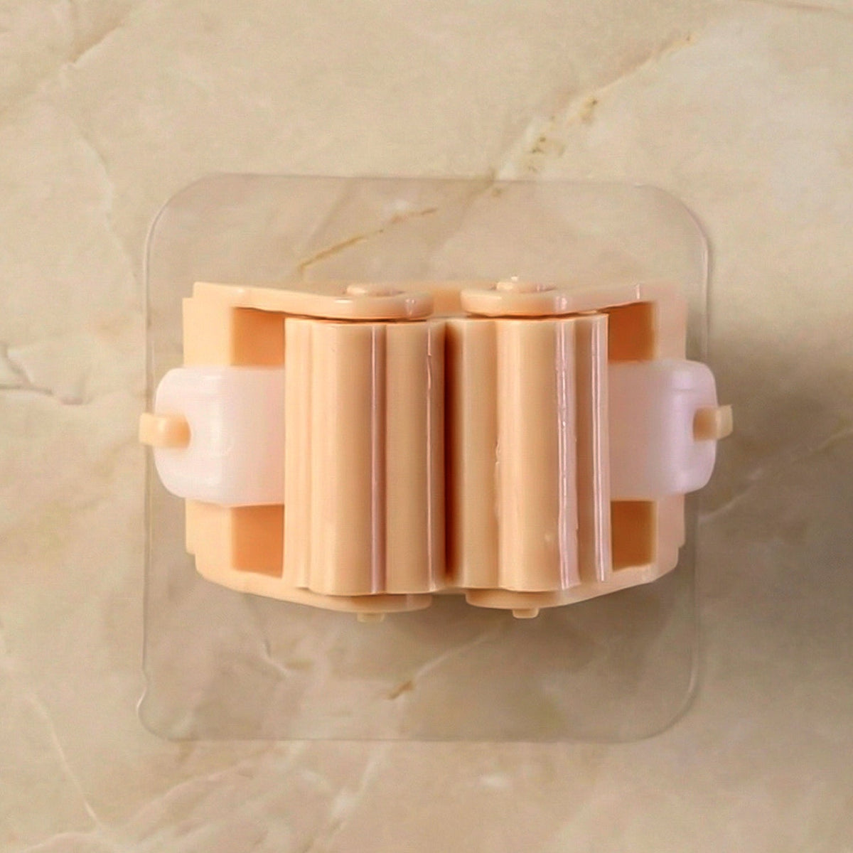 A peach-colored broom holder with white rollers is adhered to a light-colored tiled wall.