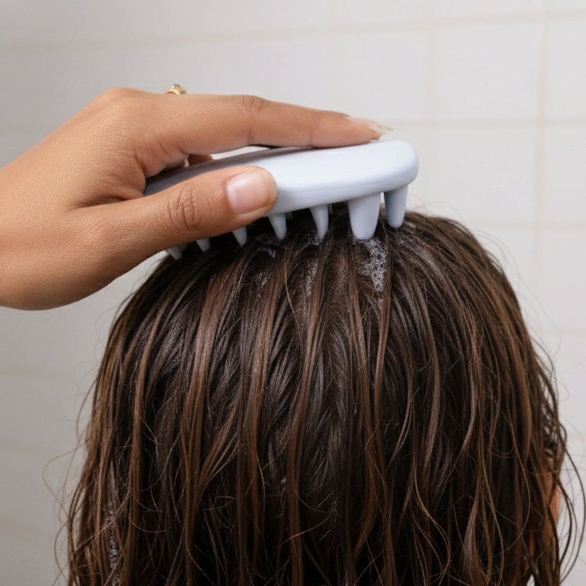A hand holding a light blue silicone scalp massager, gently massaging wet, dark hair with suds.