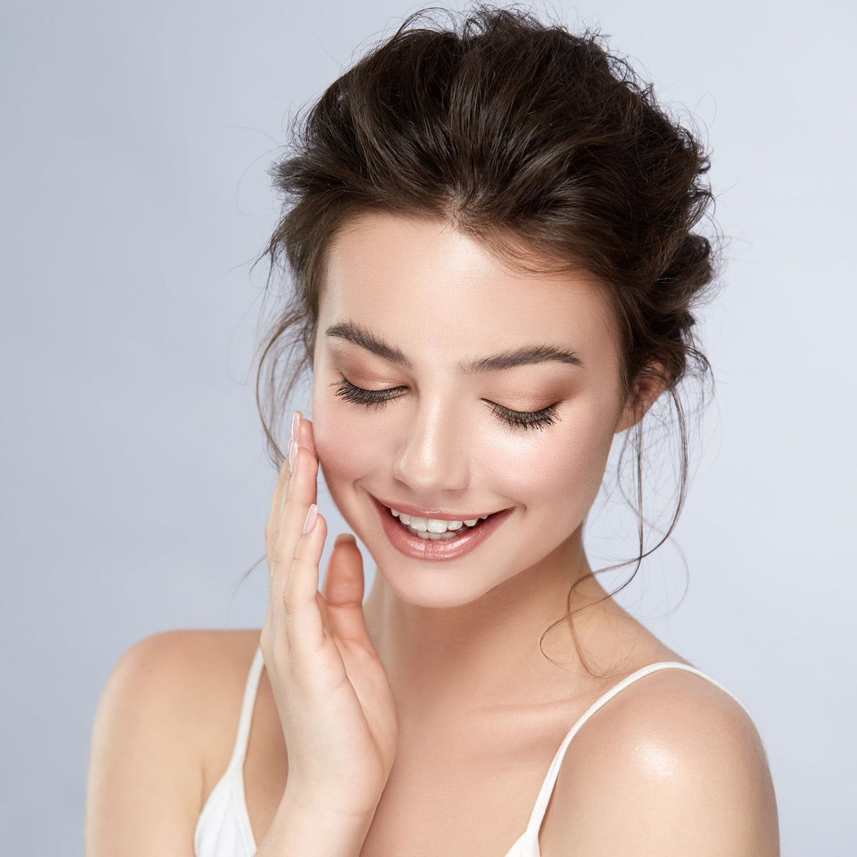 A smiling woman with dark hair and light makeup gently touches her cheek, looking down with closed eyes.