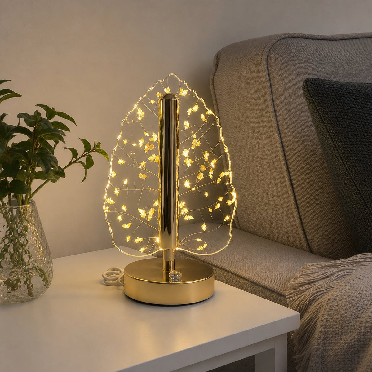 A golden crystal leaf LED table lamp on a white side table next to a green plant and a gray sofa.