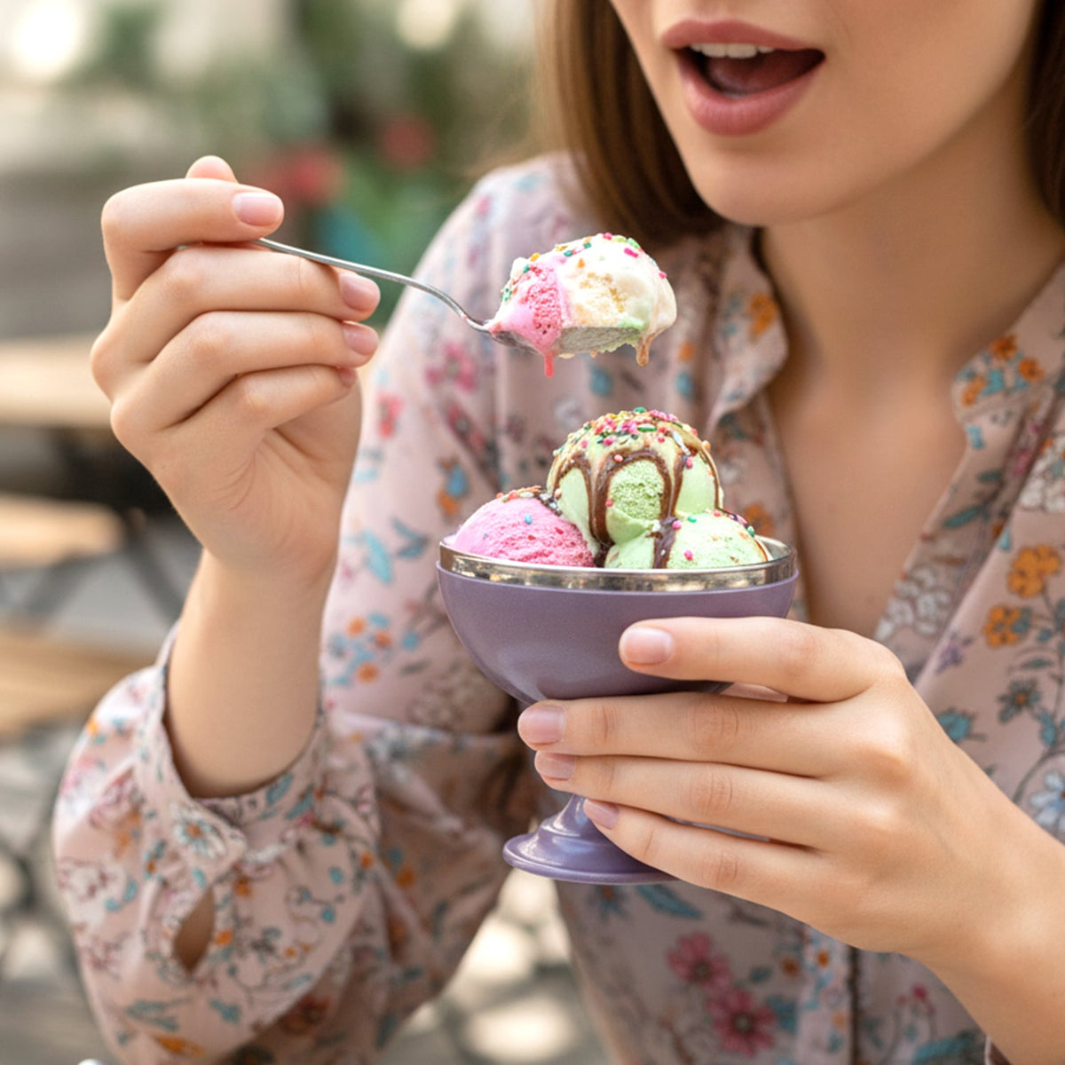 A person holds a purple dessert bowl with three scoops of ice cream, sprinkles, and sauce, about to take a bite.