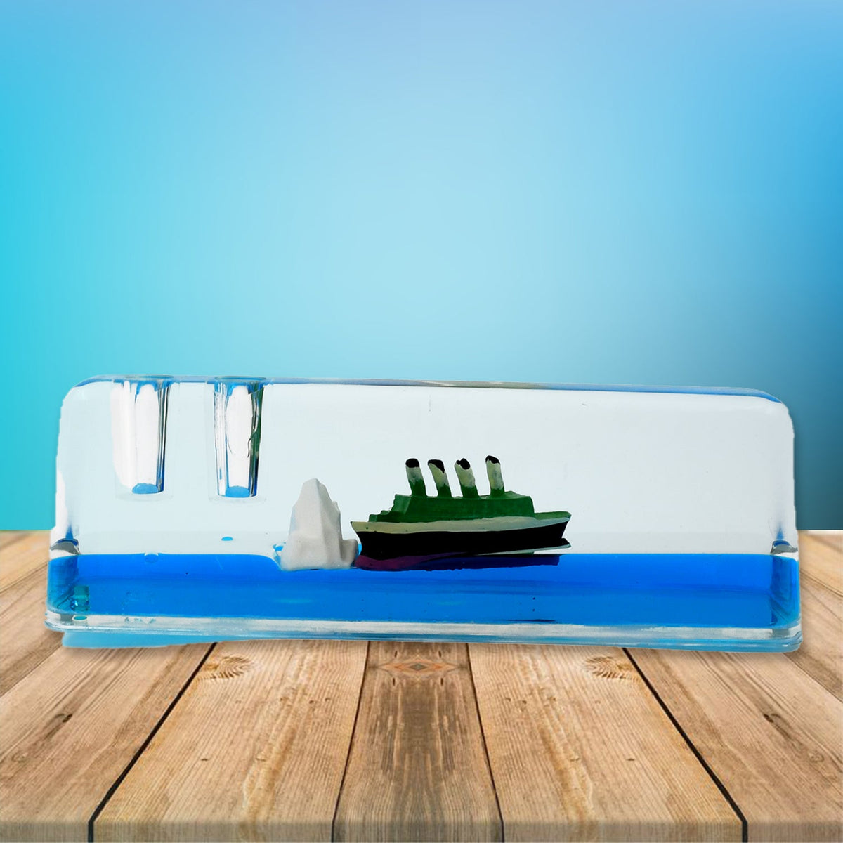 A clear rectangular liquid motion paperweight with a green ship and iceberg floats in blue liquid on a wooden surface.