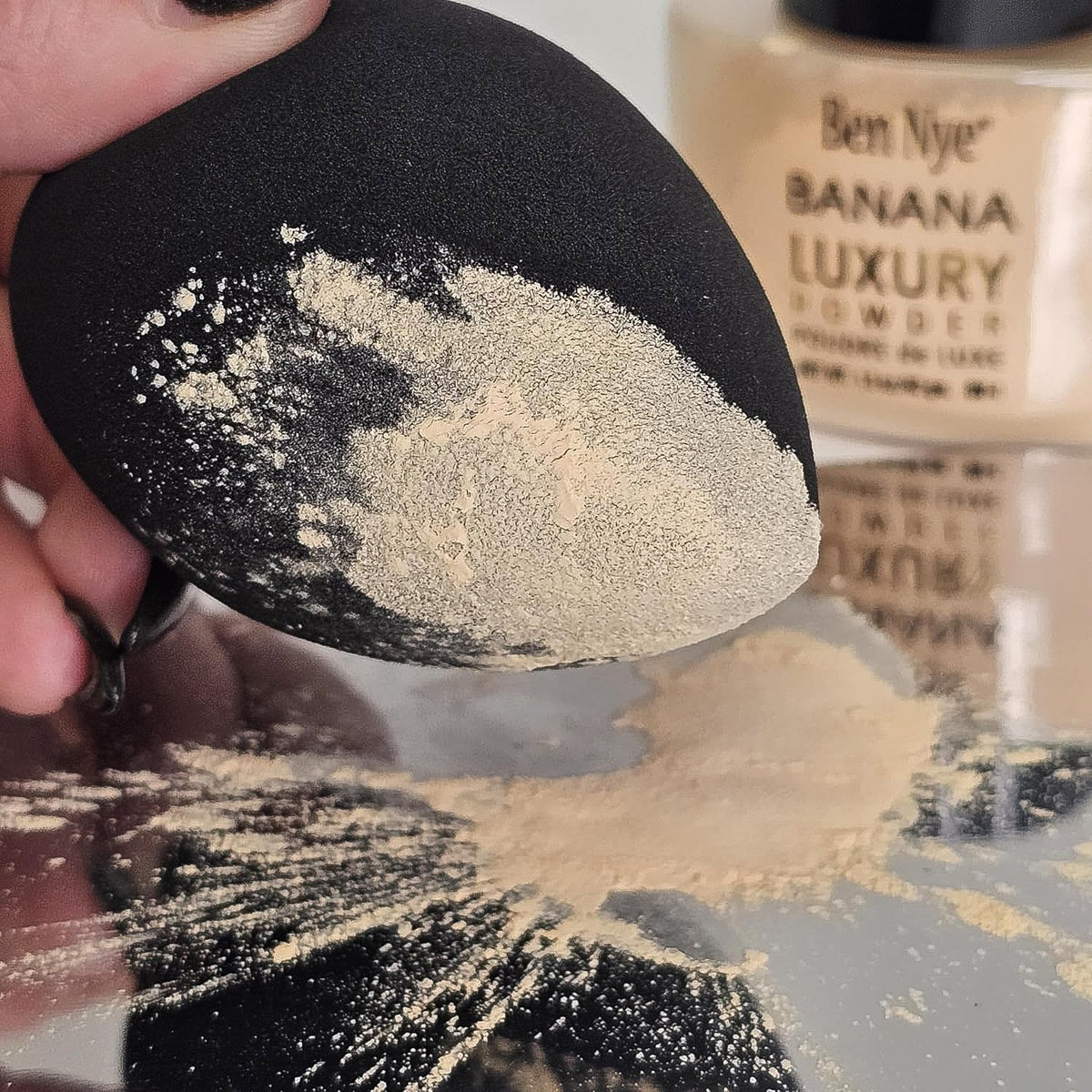 A black makeup sponge covered in light beige setting powder, with a Ben Nye Banana Luxury Powder jar in the background.