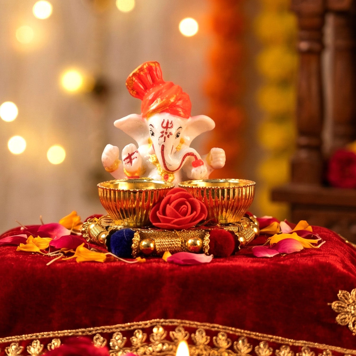 A white Ganesha idol with an orange turban sits on a gold base with two empty gold bowls and a red rose, surrounded by red