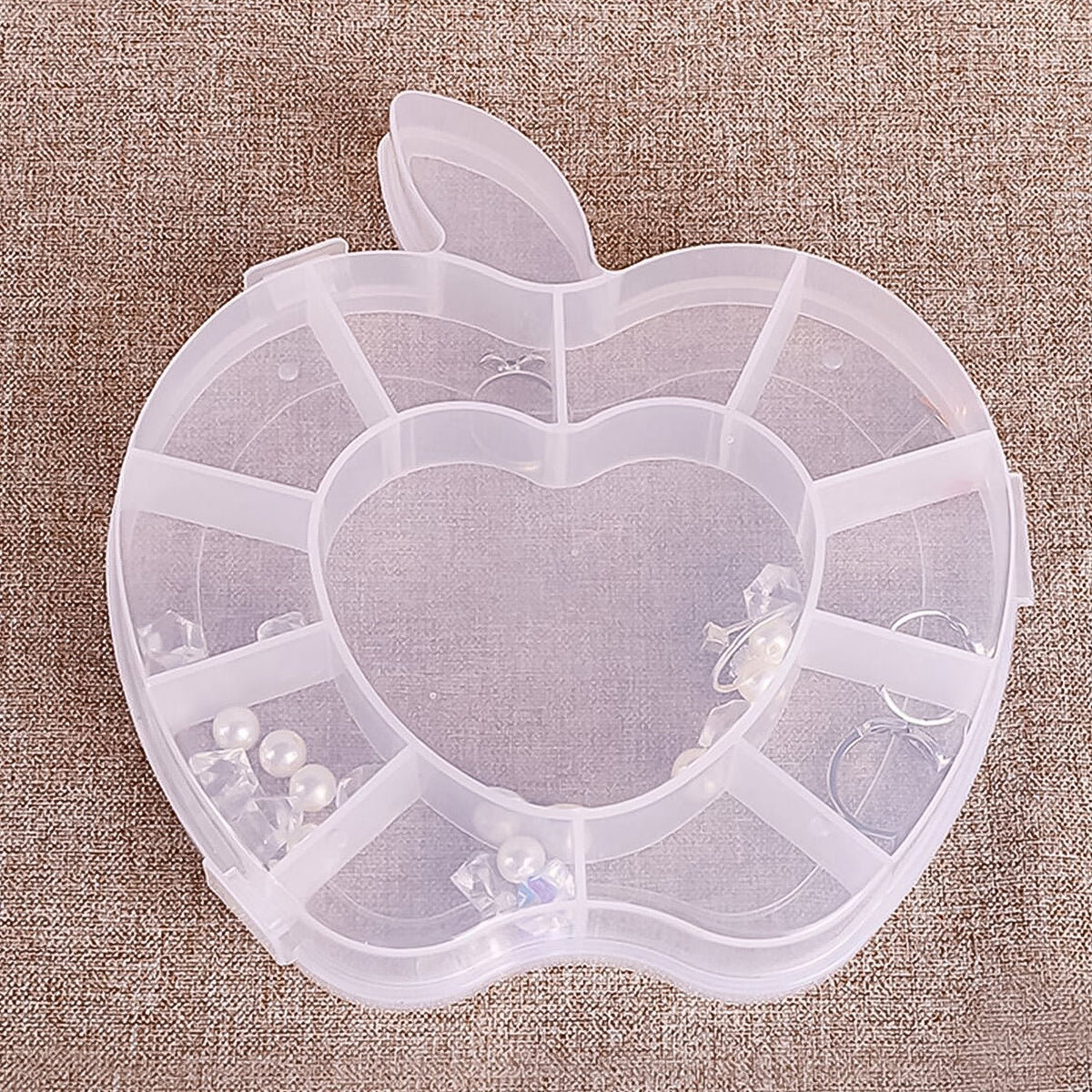 A transparent apple-shaped organizer box with multiple compartments, containing white pearls and clear beads, is placed on a