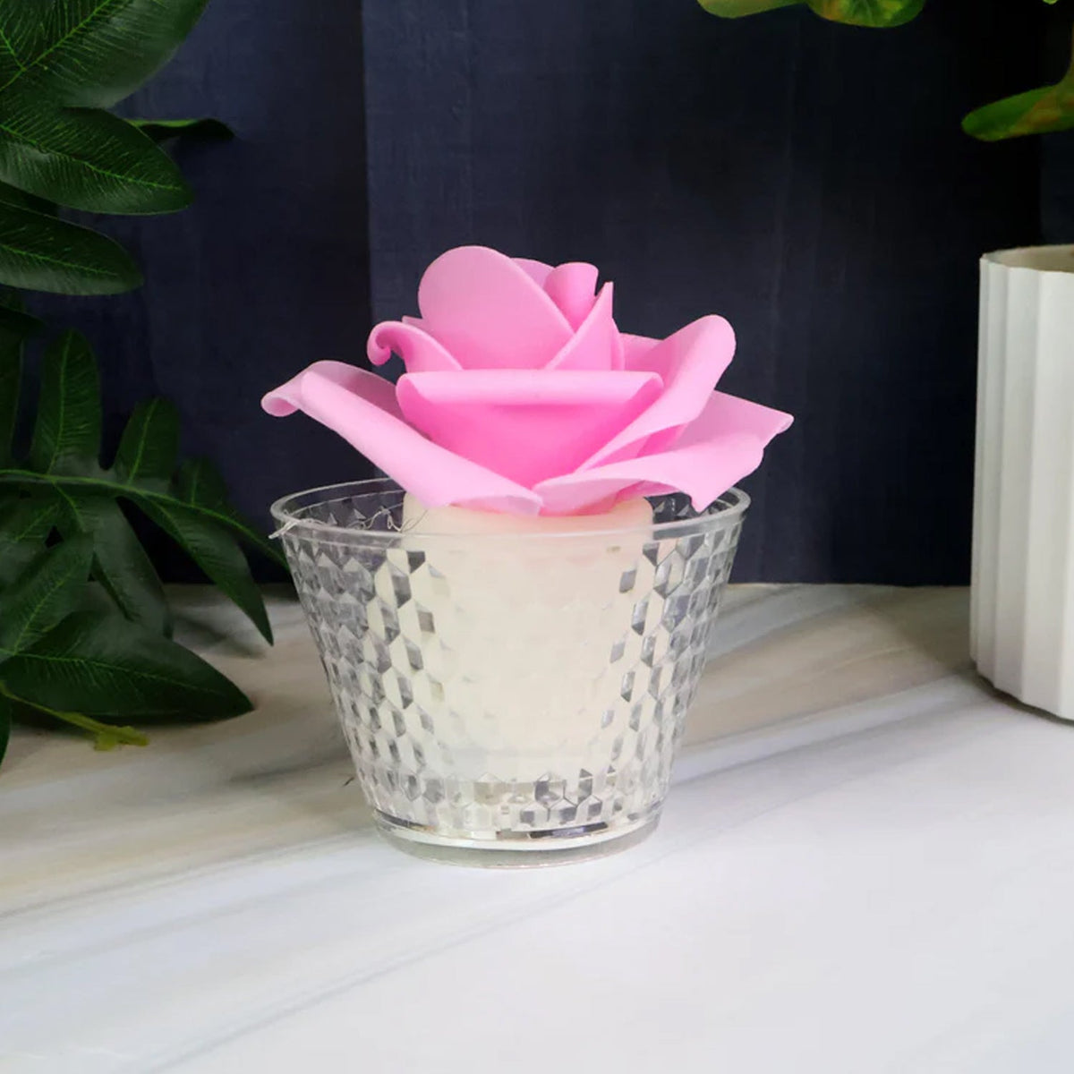 A single pink rose-shaped LED candle in a clear textured holder on a white and grey surface with green foliage.