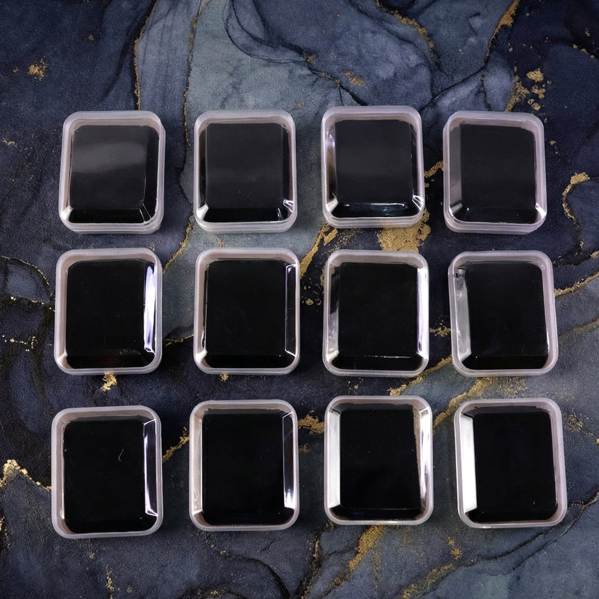 Twelve clear-lidded white jewellery boxes with black inserts arranged on a dark blue and gold marbled surface.