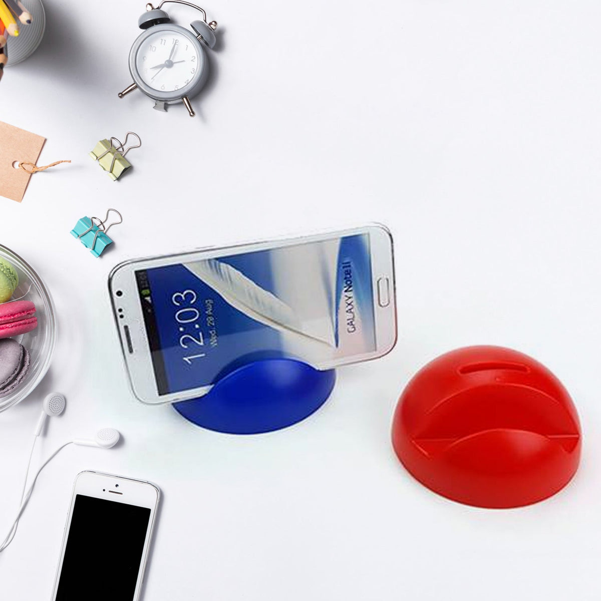 A blue phone holder with a phone and a red piggy bank on a white desk with an alarm clock and earbuds.