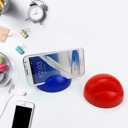 A blue phone holder with a phone and a red piggy bank on a white desk with an alarm clock and earbuds.