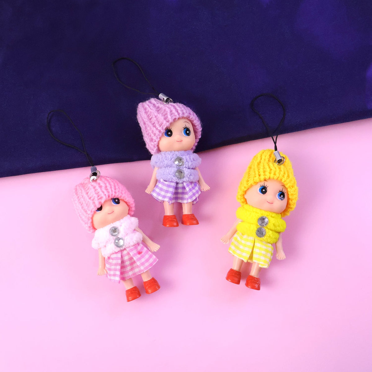Three colorful doll keychains with knitted hats stand on a pink and blue split background.