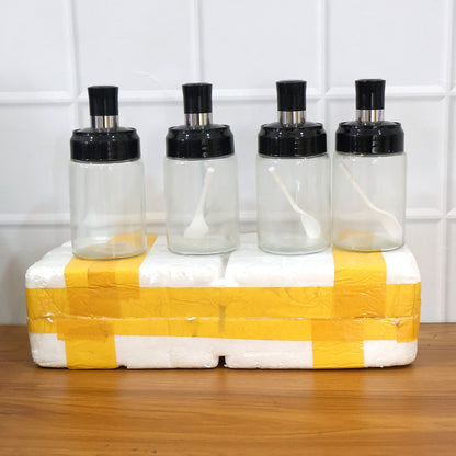 Four transparent spice jars with black lids and white spoons on a white and yellow packaged box.