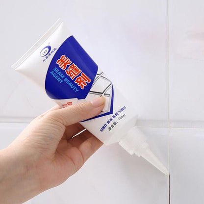 A hand holds a white and blue tube of sealant, applying it to a white tiled wall.