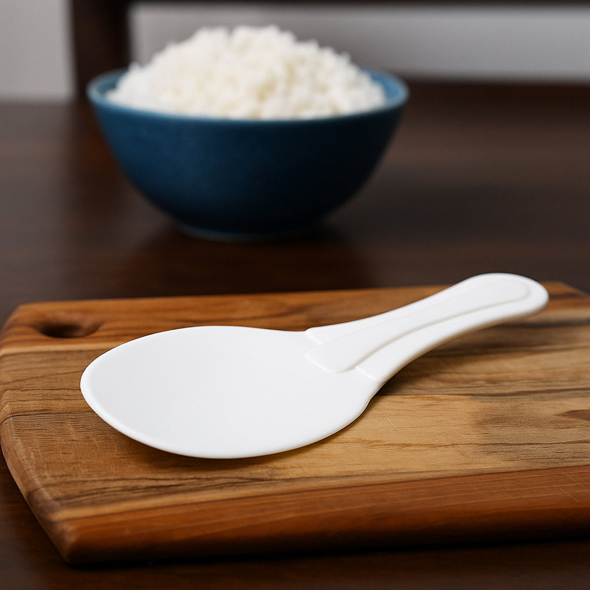 A white plastic rice paddle rests on a wooden cutting board, with a blurred blue bowl of rice in the background.