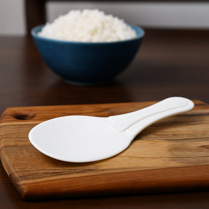 A white plastic rice paddle rests on a wooden cutting board, with a blurred blue bowl of rice in the background.