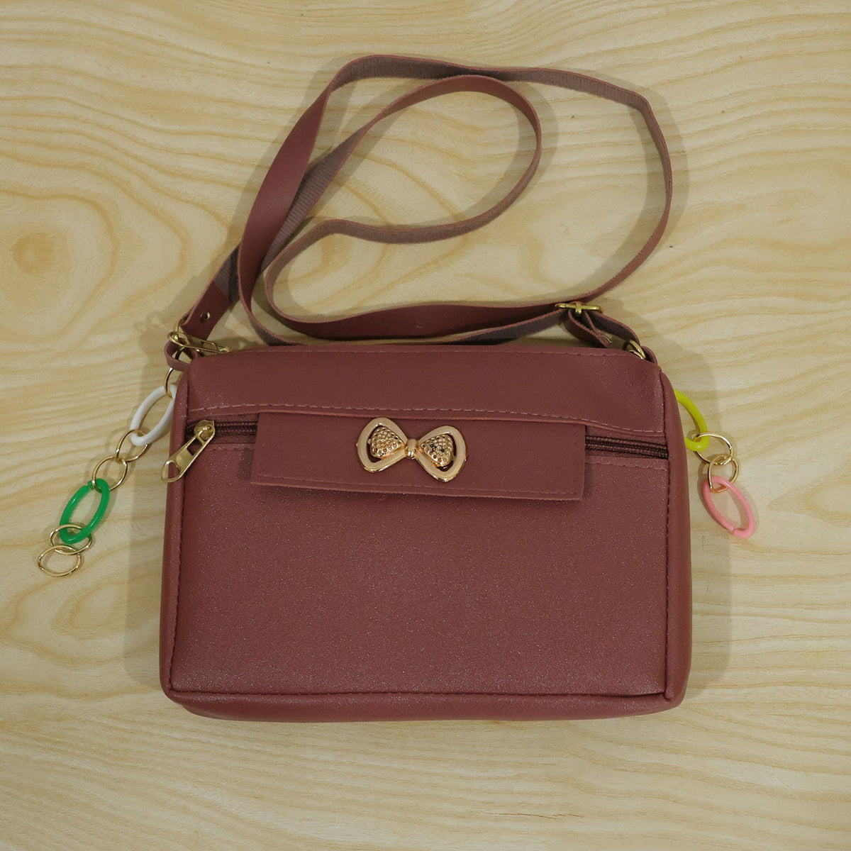 A mauve sling bag with a gold bow and colorful charms, laid flat on a light wooden surface.