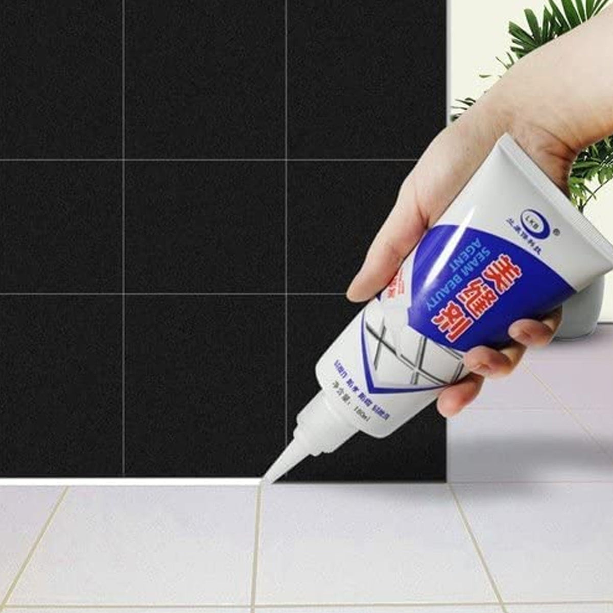 A hand applies white sealant from a tube to the grout line between white floor tiles and black wall tiles.
