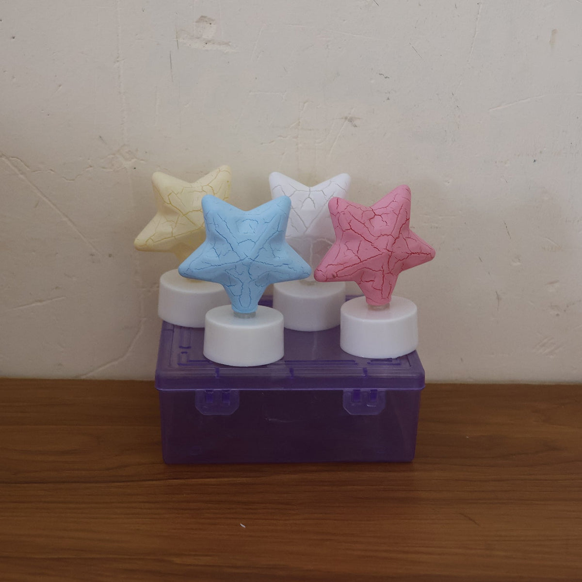 Four unlit star-shaped LED tealight candles in yellow, blue, white, and pink, displayed on a purple plastic box.