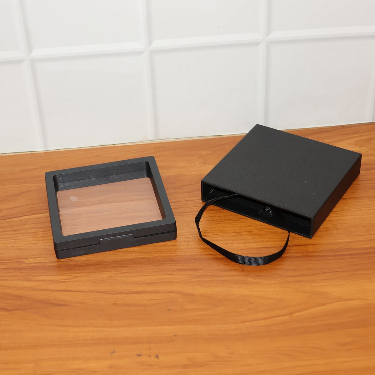 Two black square box pieces, one with a clear film and a wooden base, the other with a black ribbon.