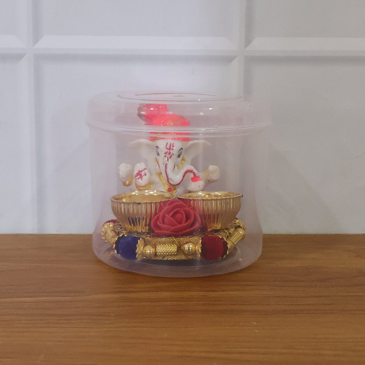A white Ganesha idol with an orange turban on a gold base with two empty gold bowls and a red rose, enclosed in a clear
