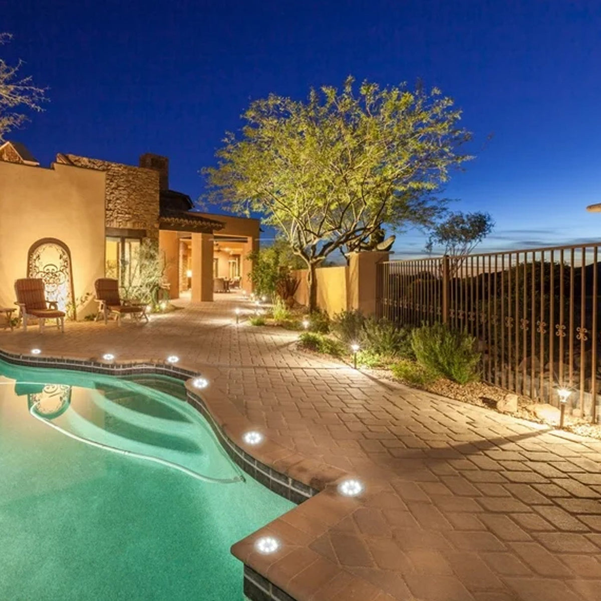 A luxurious outdoor area with a swimming pool and paved patio is illuminated by numerous small ground lights at twilight.