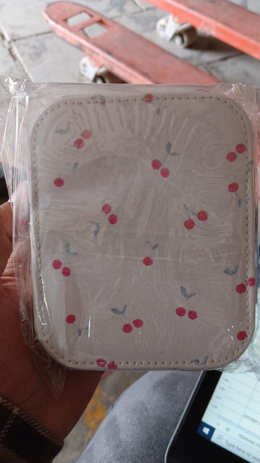 A hand holds a cherry print jewelry organizer, still in its clear plastic packaging, above a blurred background.