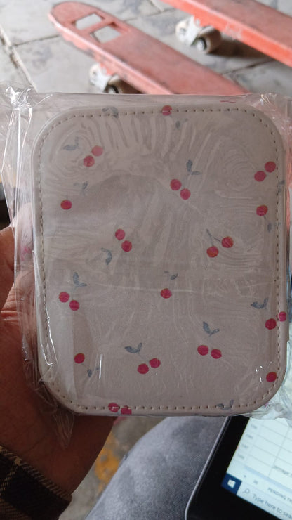 A hand holds a cherry print jewelry organizer, still in its clear plastic packaging, above a blurred background.