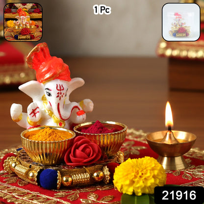 A white Ganesha idol with an orange turban on a gold base with two bowls, one filled with yellow powder and the other with