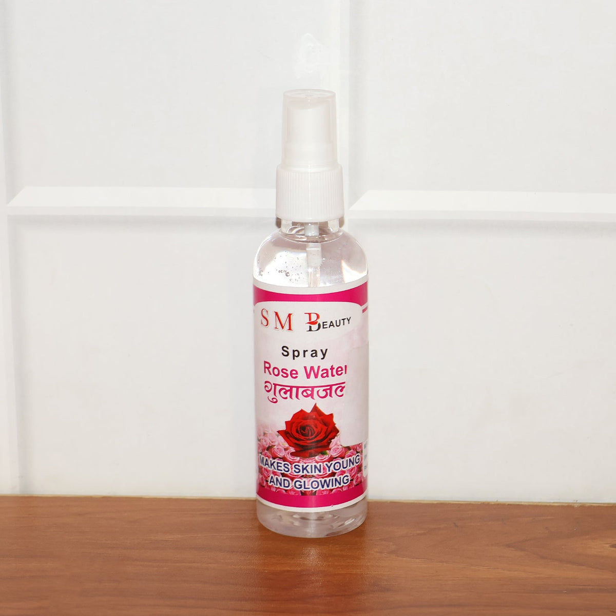 A clear bottle of SM Beauty rose water spray with a pink label stands on a wooden surface against a white tiled wall.