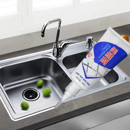 A hand applies white sealant from a tube to the edge of a stainless steel kitchen sink with green limes.