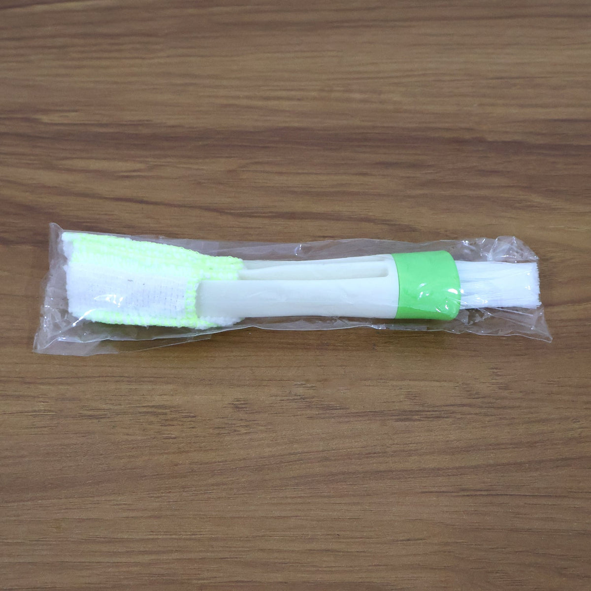 A white and green 3-in-1 cleaning brush sealed in clear plastic packaging on a wooden surface.