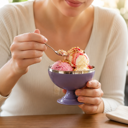 A person holds a purple dessert bowl filled with two scoops of ice cream, topped with sauce and nuts.
