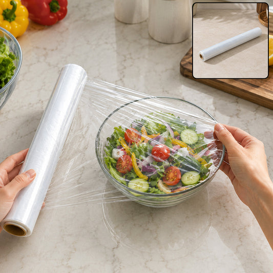 Hands wrapping a glass bowl of salad with clear plastic film on a marble counter, with a cling wrap roll beside it.