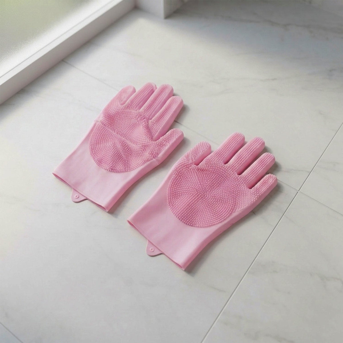 A pair of pink silicone scrubber gloves lying on a light-colored tiled floor near a window.