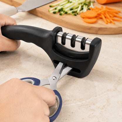Hands hold a black three-stage sharpener, sharpening blue-handled scissors with chopped vegetables in the background.
