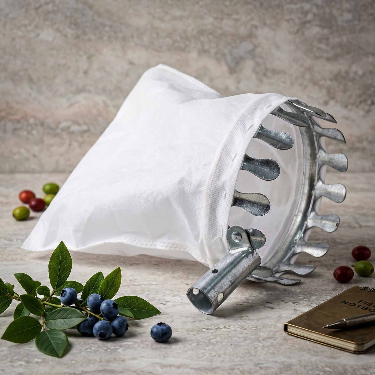 The fruit picker's white bag and metal prongs are on a textured grey surface, surrounded by blueberries, leaves, and a