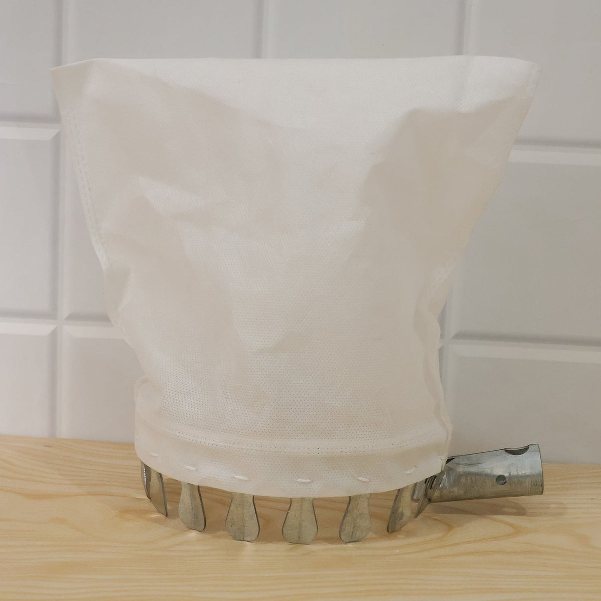 The white collection bag and metal prongs of the fruit picker are on a light wooden surface against a white tiled background.