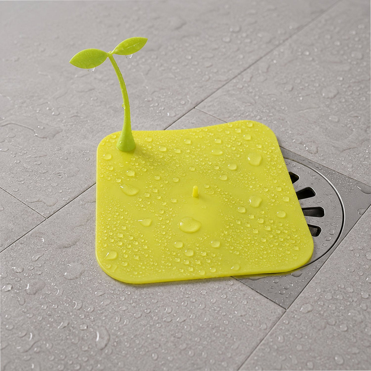 A bright yellow square silicone drain cover with a green sprout-like handle, covered in water droplets on a wet floor.