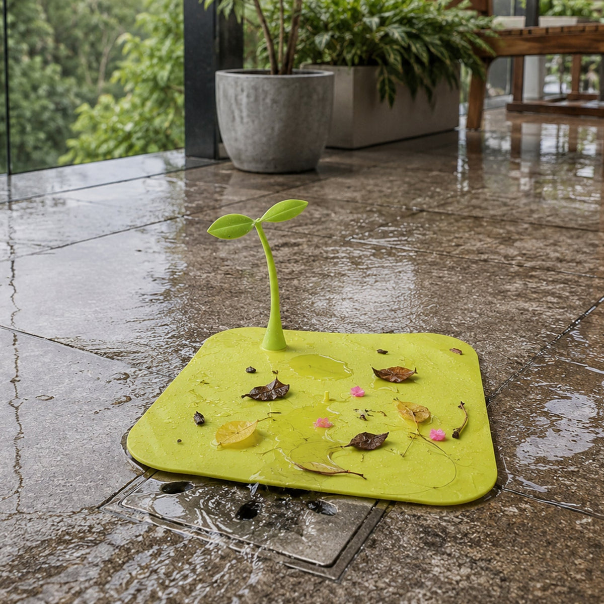 A bright yellow square silicone drain cover with a green sprout-like handle, collecting leaves and debris on a wet patio.