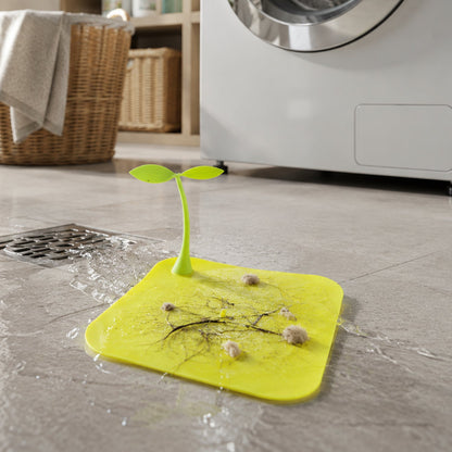 A bright yellow square silicone drain cover with a green sprout-like handle, collecting lint and hair on a laundry room