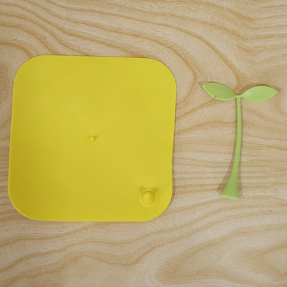 A bright yellow square silicone drain cover and its detached green sprout-like handle on a light wooden surface.
