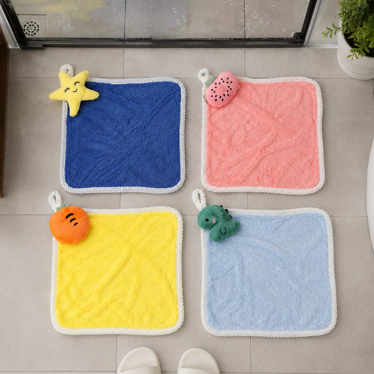 Four colorful square towels with animal and fruit shapes laid flat on a light grey tiled floor.