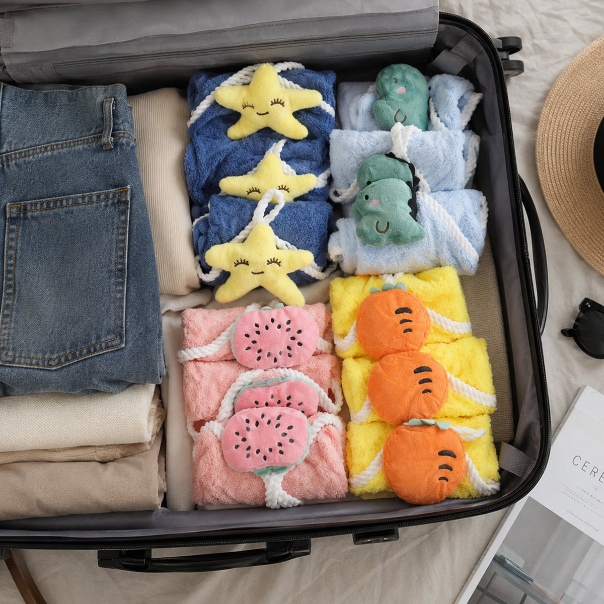 An open suitcase filled with neatly packed clothes and twelve colorful towels with plush animal and fruit shapes.