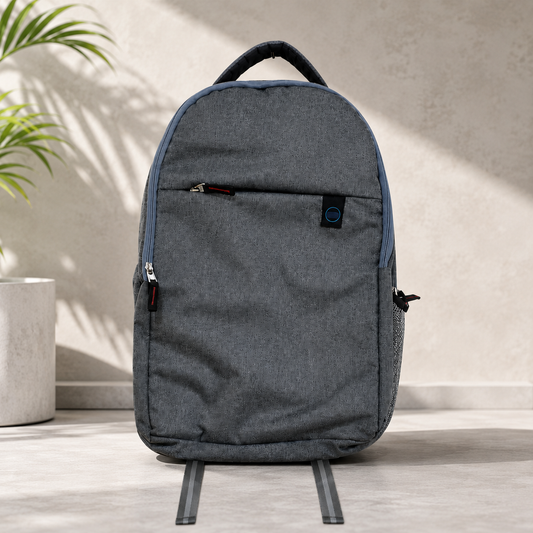A grey backpack with a front zipper pocket and blue trim stands on a light grey surface next to a plant.