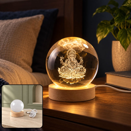A clear crystal sphere with a detailed Ganesha engraving sits on a wooden LED base, glowing warmly on a bedside table.