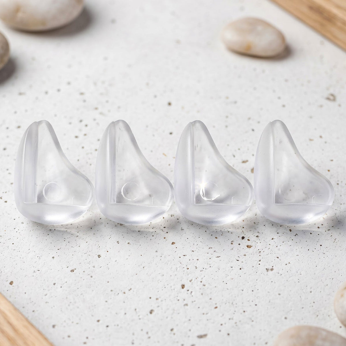 Four clear, rounded silicone corner protectors are lined up on a speckled white surface with light pebbles.