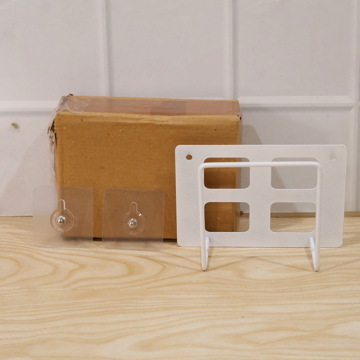 A white metal router mount, two clear adhesive hooks, and a brown cardboard box on a light wooden surface.
