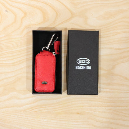 Red leather car key case with a silver snap hook, placed inside a black gift box next to a branded black box.