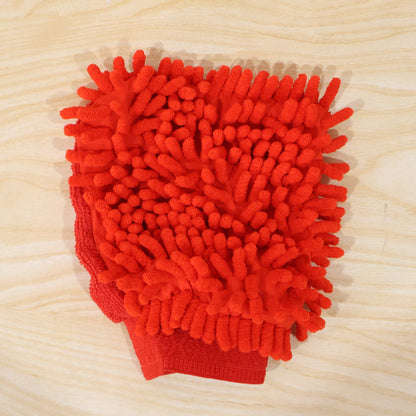 A red microfiber chenille wash glove with a textured surface lies flat on a light wooden surface.