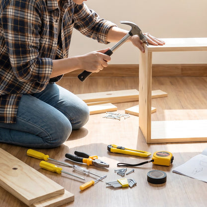 A person in a plaid shirt and jeans kneels on a wooden floor, using a hammer to assemble a light wood frame.